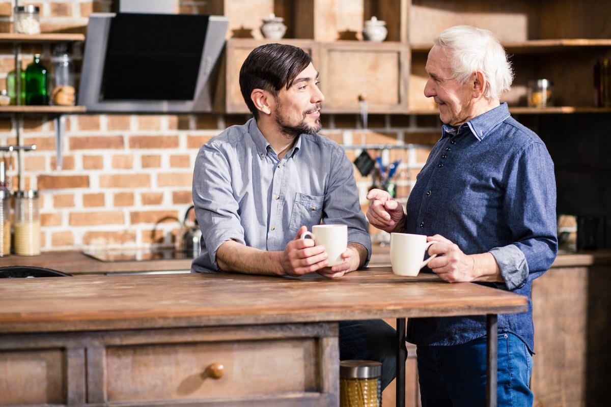 Two men having a friendly conversation over coffee in a modern kitchen, discussing dental treatment options.