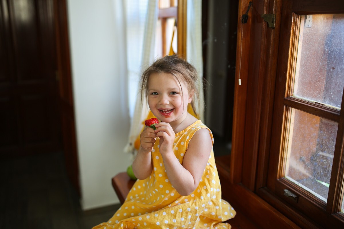 Best Summer Foods for Your Teeth Happy child in yellow polka dot dress holding fresh strawberry indoors.