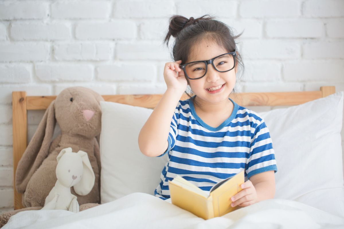 Young girl wearing glasses reading a book with a stuffed bunny, smiling brightly.
