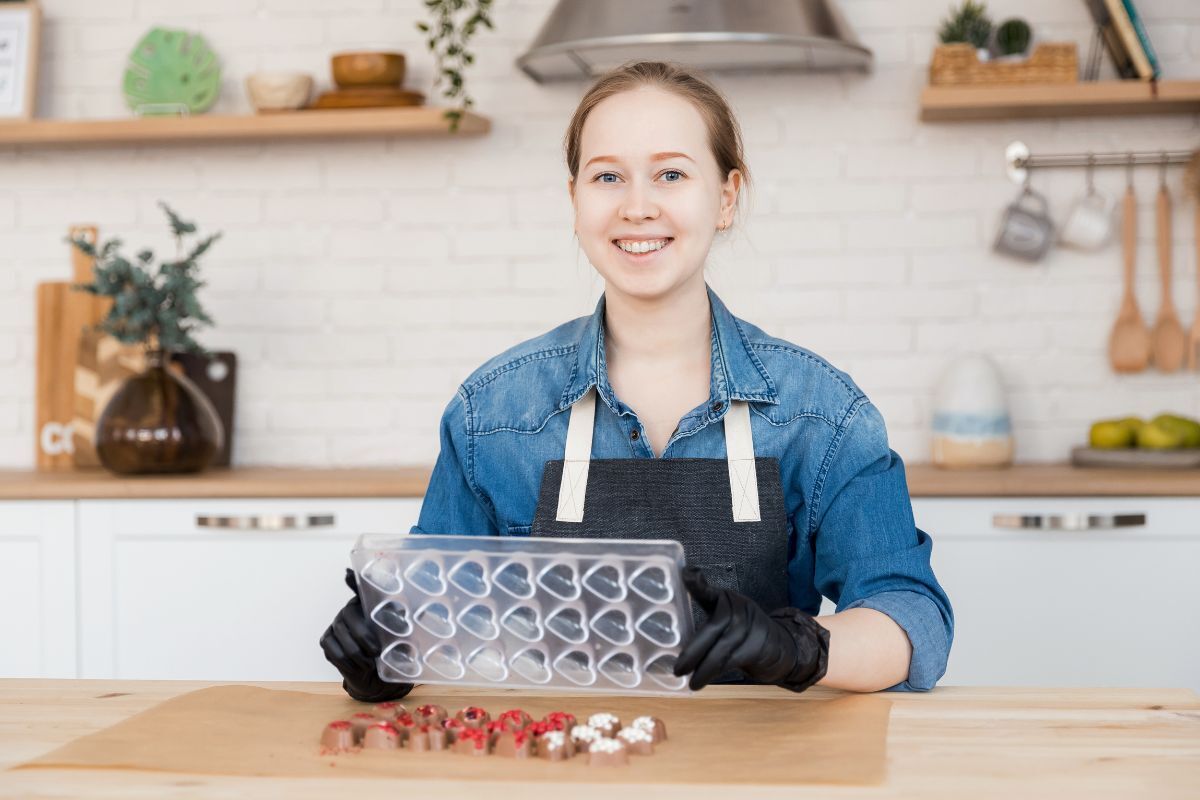 Valentines Day Candy Shopping Tips Young woman in denim shirt holding heart-shaped candy mold in modern kitchen setting.