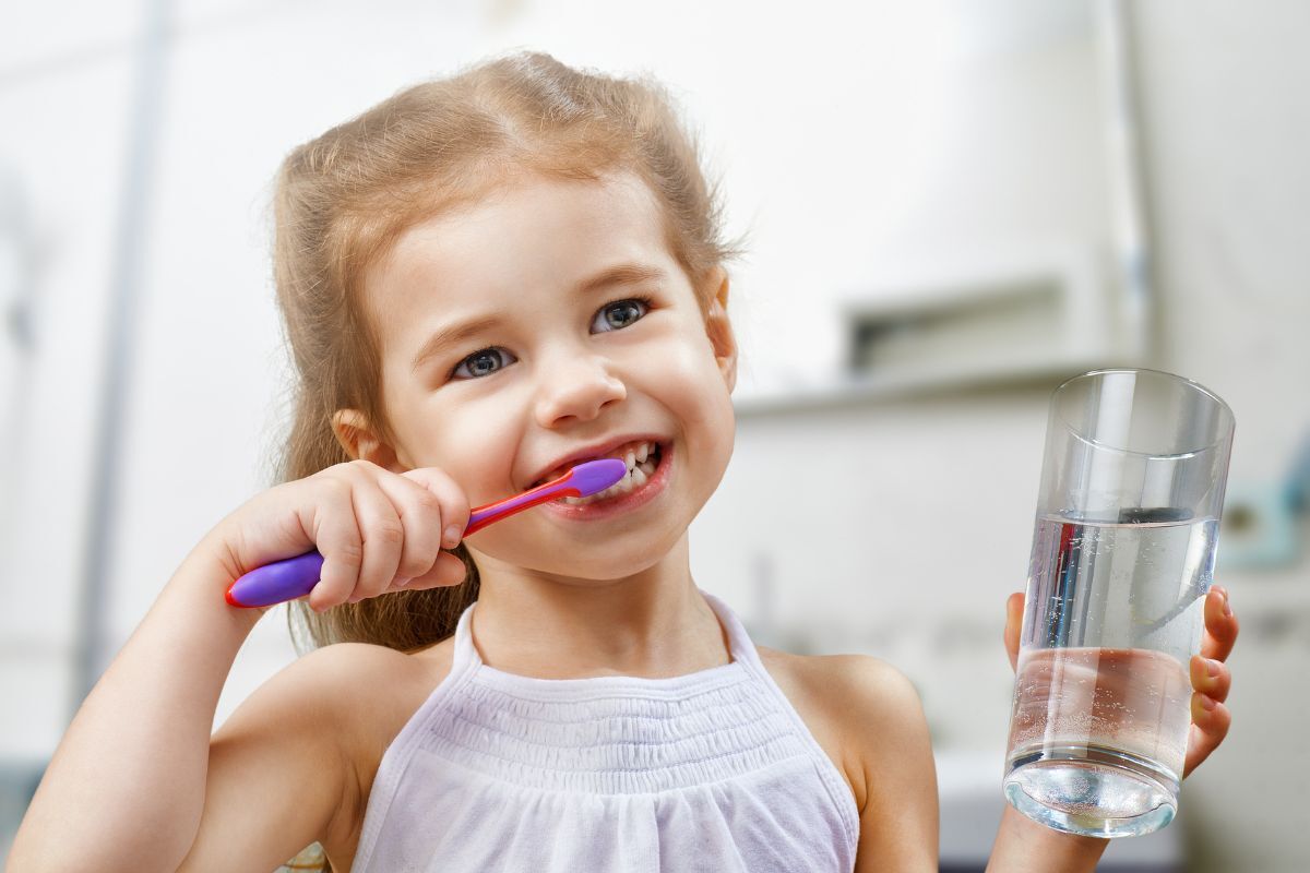 Happy young girl brushing her teeth with a colorful toothbrush and holding a glass of water.